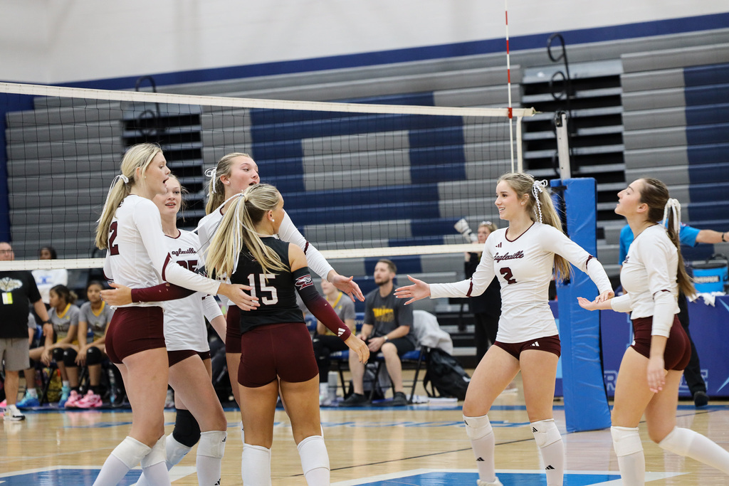Indoor volleyball court. Girls in white and maroon jerseys celebrating, standing around a net. Spectators in the background.