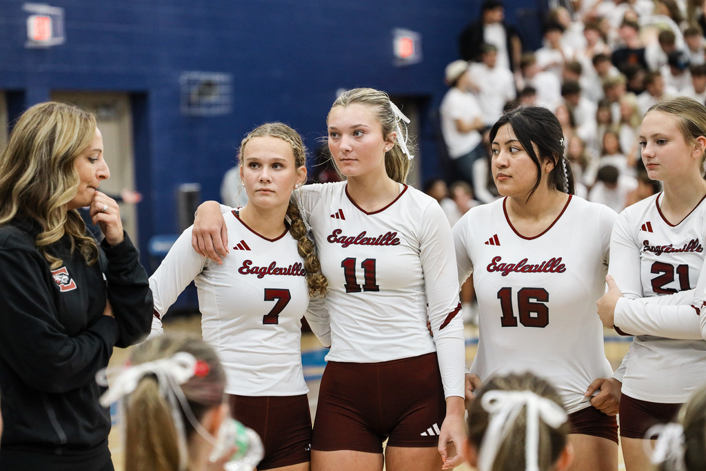 Five female volleyball players in white and maroon uniforms stand together, facing a coach.