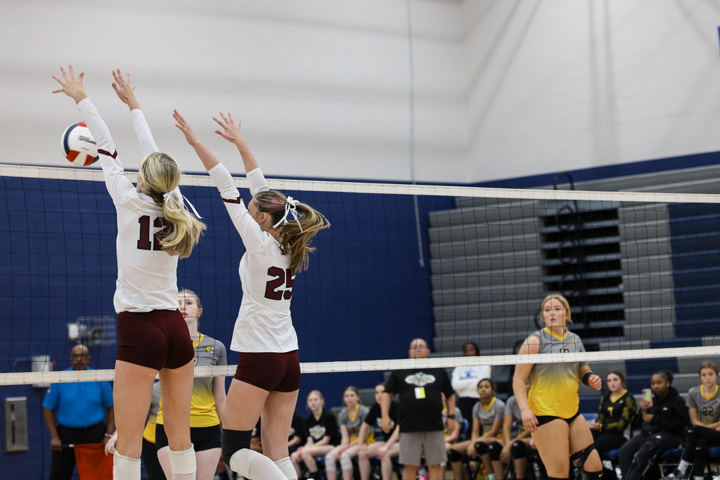 Two women play volleyball, one jumping to block the ball, the other reaching to hit it.