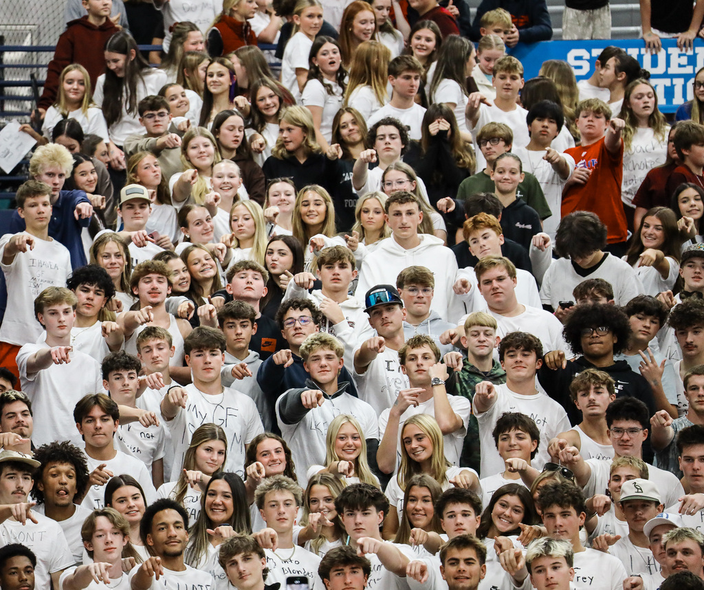 A large crowd of students wearing white shirts, standing in a stadium with raised hands.
