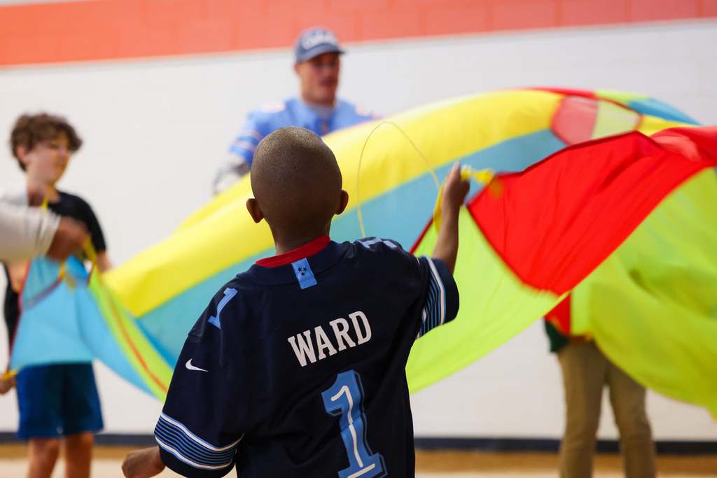 Boy with jersey number 1 holding strings of a large kite, surrounded by other children and adults.