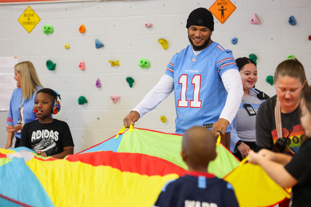 People in a gym. A man in a blue jersey holds a colorful parachute. Two children play under it.