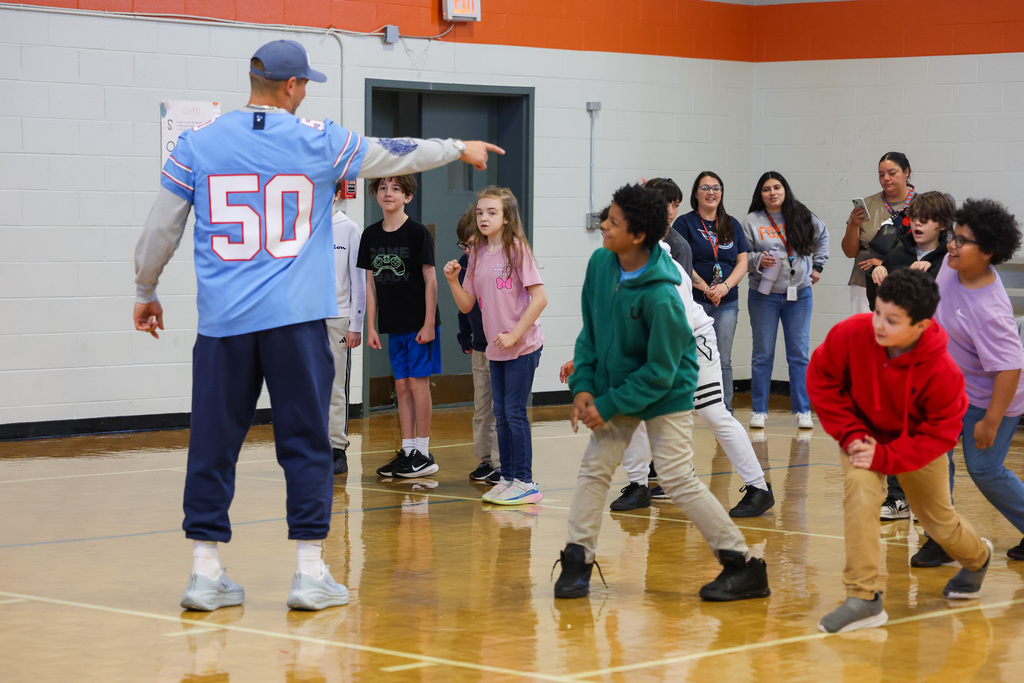 A man in a blue jersey points inside a gym. Children in jerseys and sneakers move about.