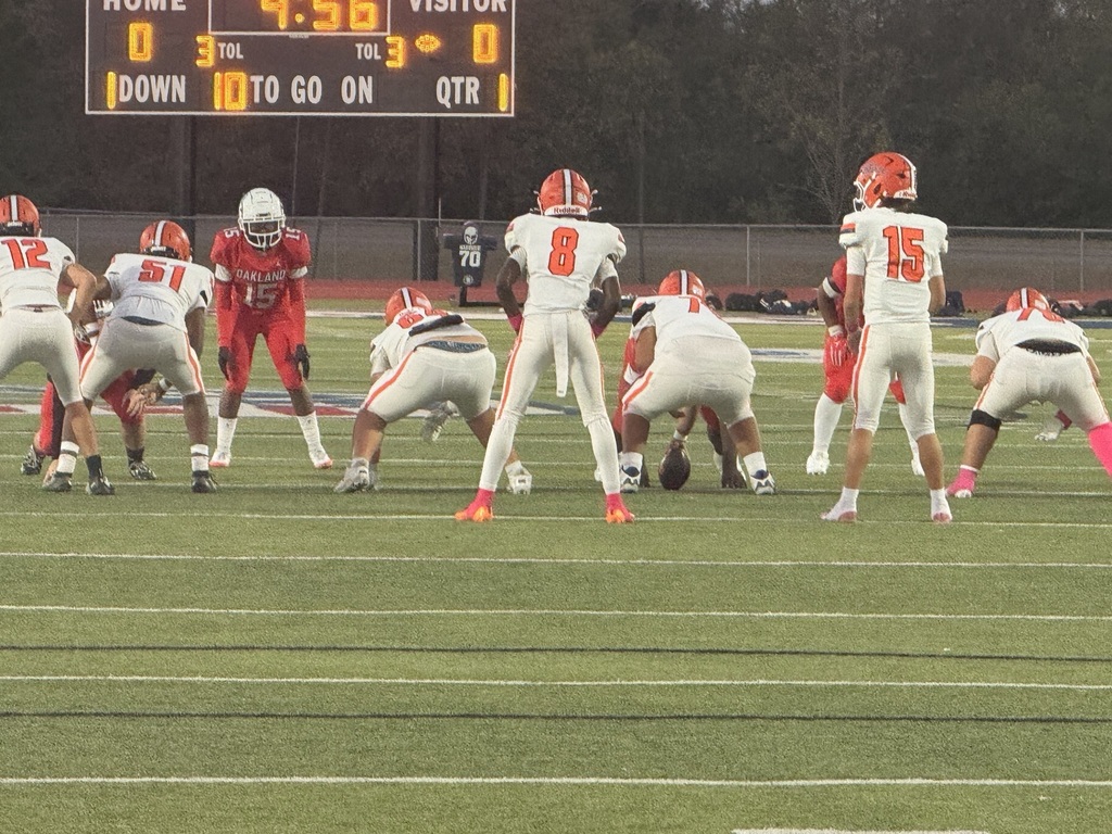 Football players wearing uniforms, helmets, and sneakers on a field. A scoreboard above showing scores.
