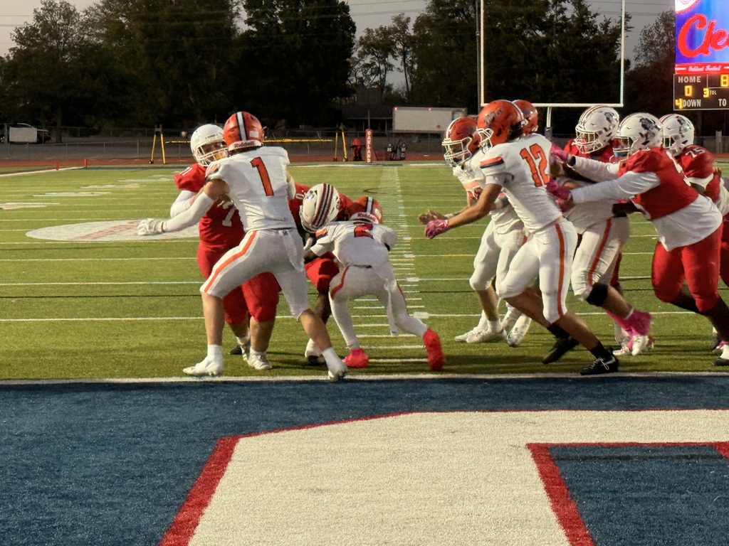 Football players wearing uniforms, helmets, and sneakers on a field. A scoreboard above showing scores.