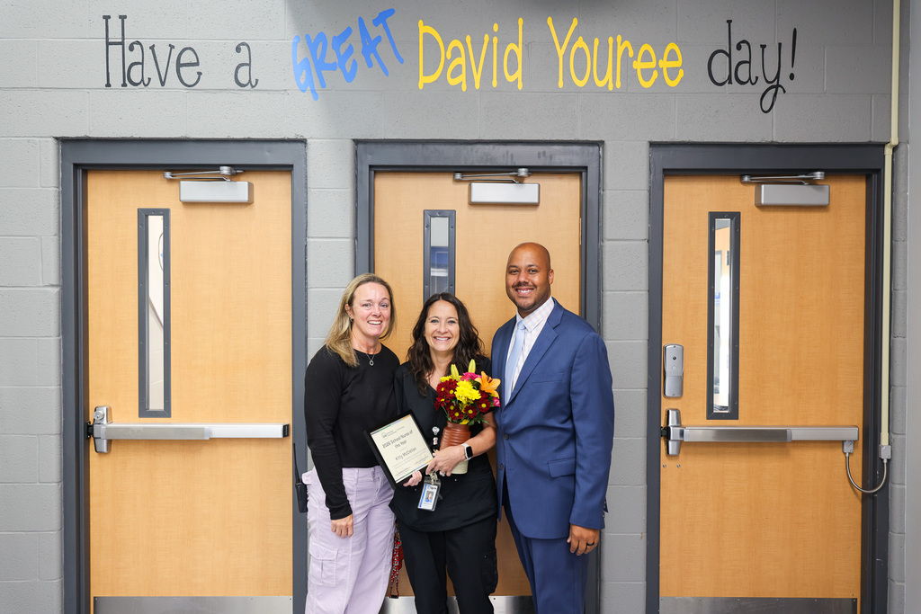 Three people stand before three closed doors, one holding a bouquet of flowers, another holding a certificate.