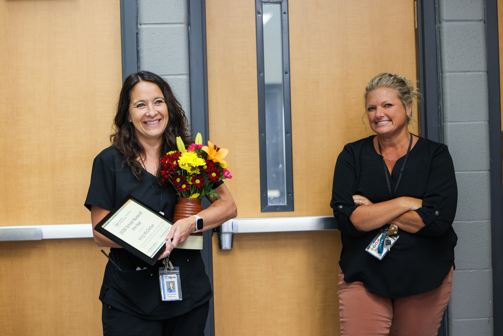 Two women smile for a photo. One holds a framed certificate and flower arrangement. Both wear ID badges.