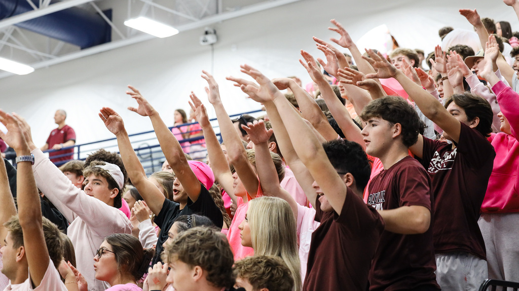 Group of students raising hands, many wearing maroon and pink, some wearing caps. Brightly lit gym with bleachers.