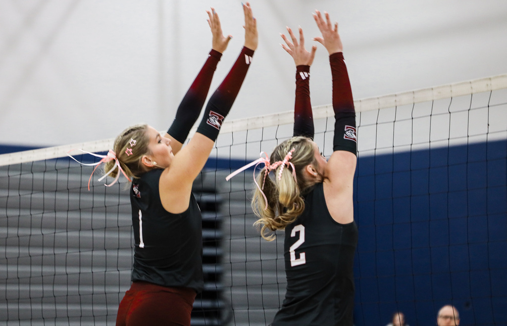 Two women jumping to block volleyball over net. One wears jersey with number 1, other with 2.