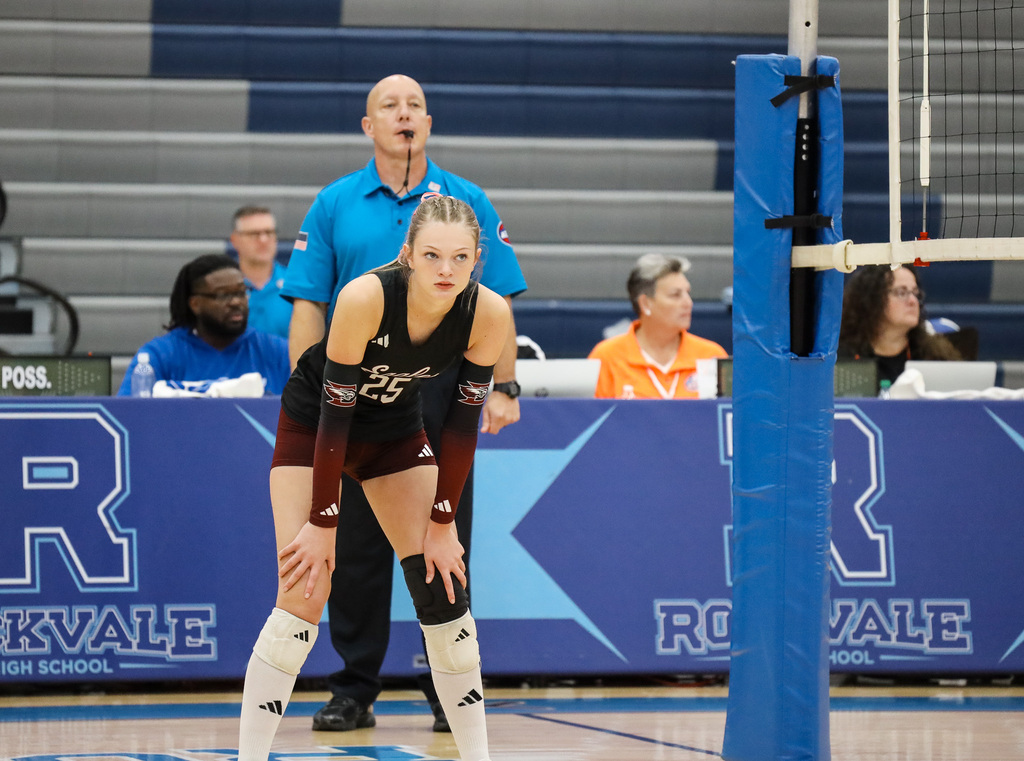 Volleyball player in uniform crouches on indoor court, with referee and spectators in background.
