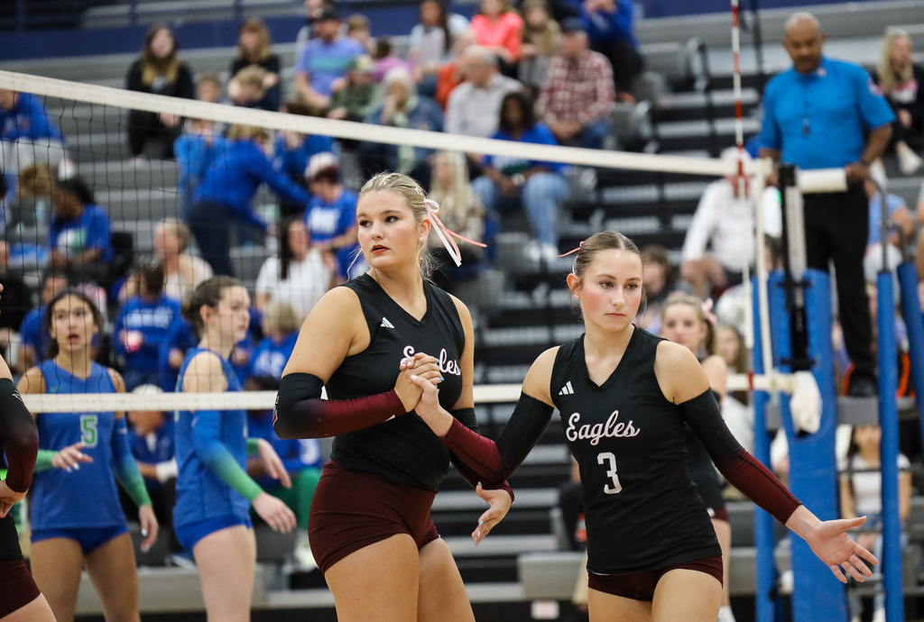 Two women in black and red uniforms with white numbers stand on a court. A crowd watches from bleachers.