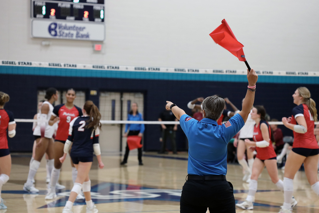 An indoor gym with a scoreboard, a group of women wearing red and blue uniforms, a man holding a flag.