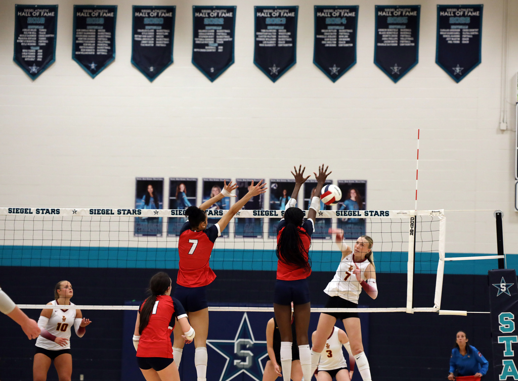 Players in numbered jerseys jumping, hands raised, in front of volleyball net. One player holding a ball.