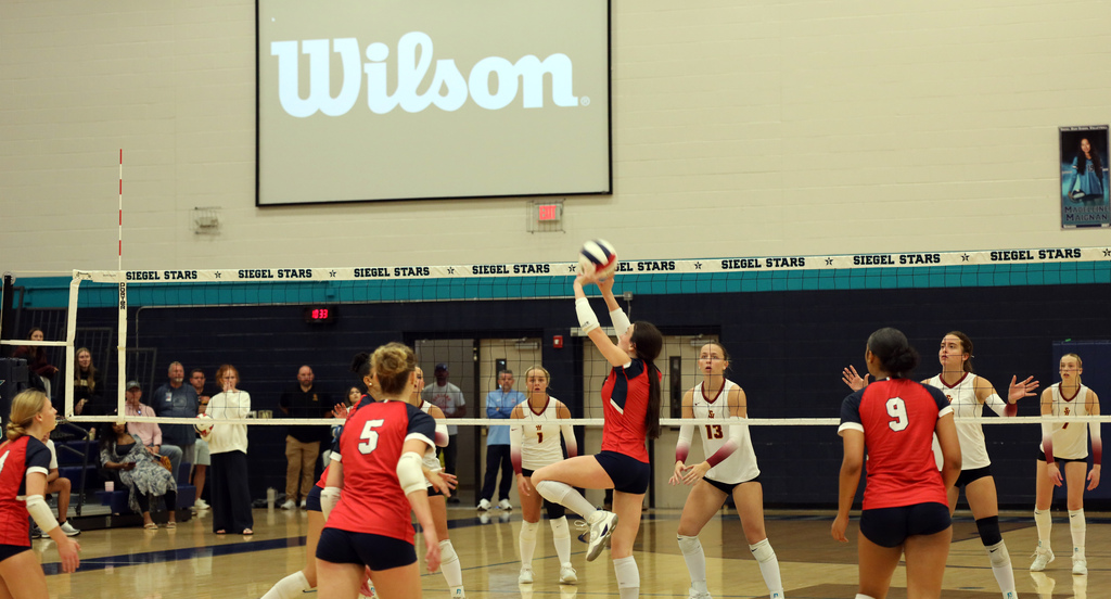 Wilson banner on wall above volleyball players. Player in red jersey jumps and hits ball. Two players in white jerseys block.