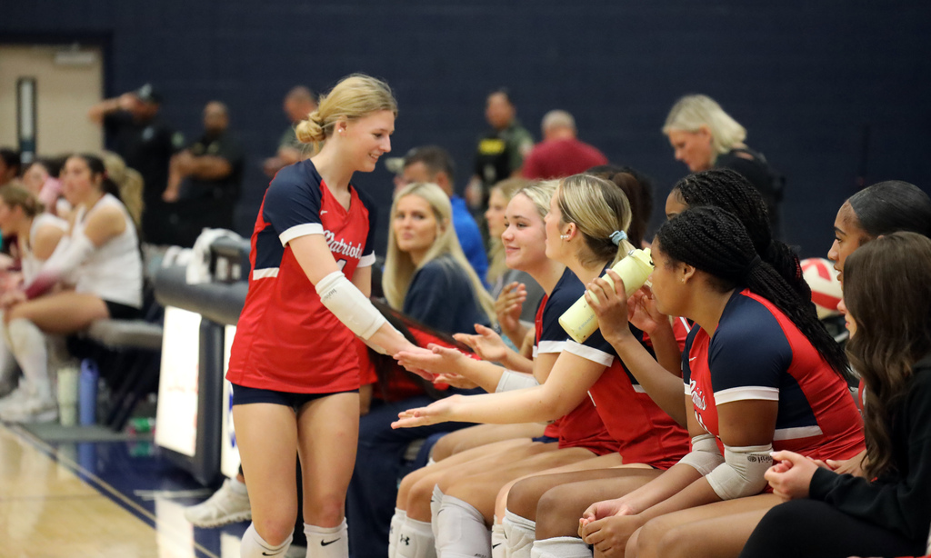A group of young women in matching uniforms sit and stand on a gym bench and court.