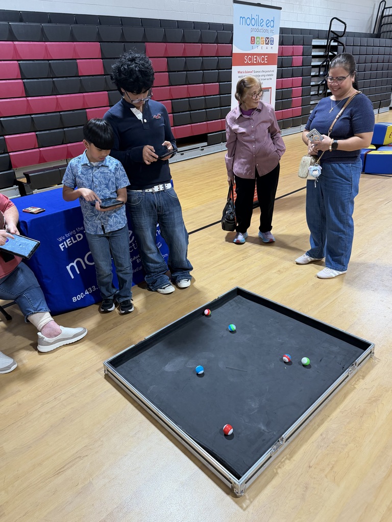 Group of adults and children at a school event, watching people play a game with balls on a game board.