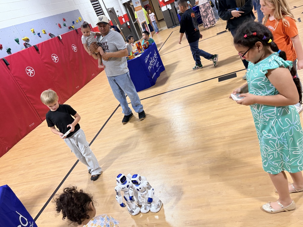 Group of children and adults in a gym with a blue banner. Some children play with robots.