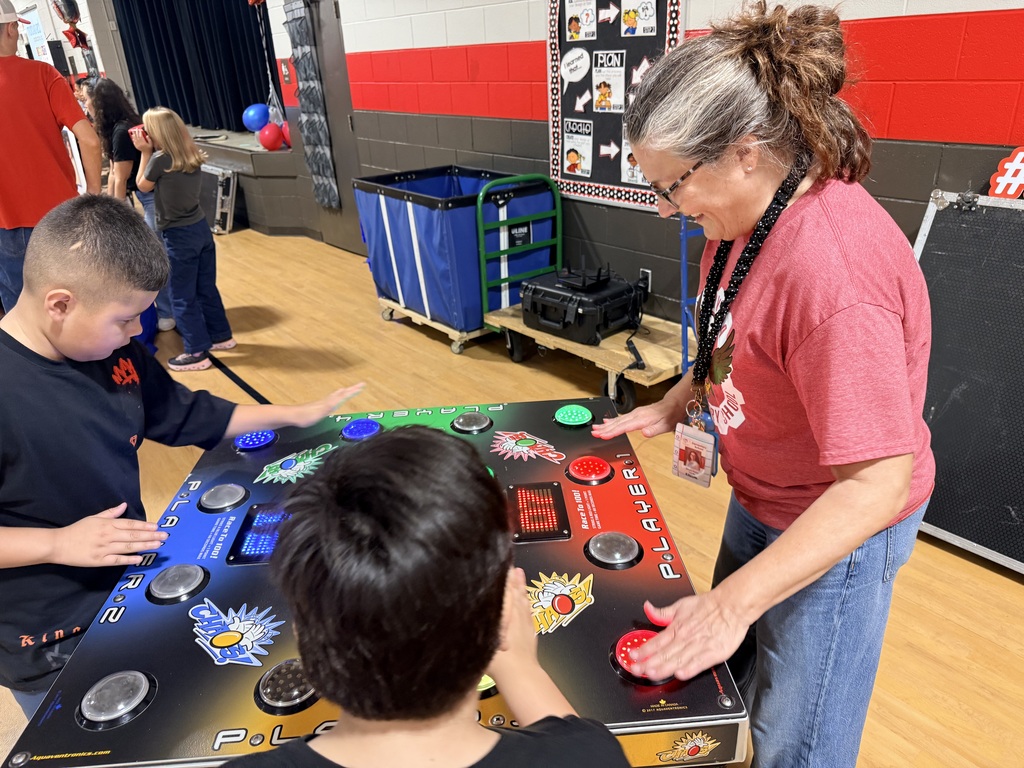 Two children play an arcade game while a woman in glasses and a pink shirt helps them.