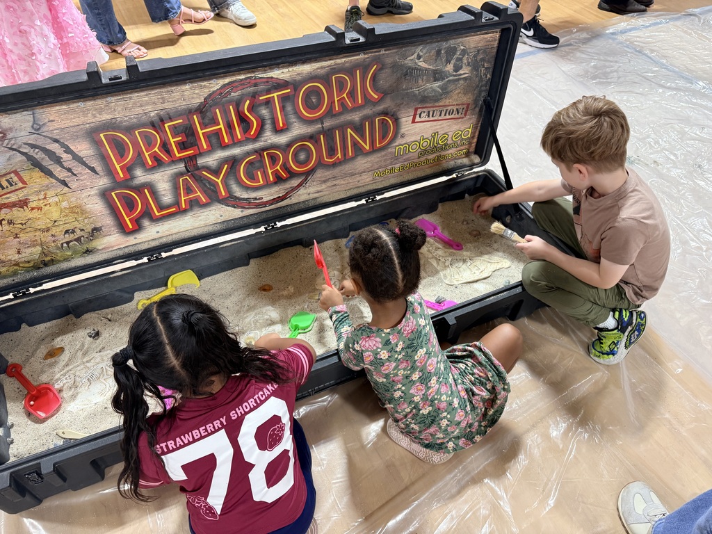 Three children play in a sandbox labeled "Prehistoric Playground," with one using a red shovel.