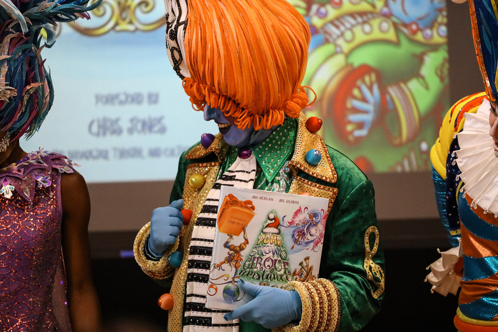 Two people in colorful costumes, one holding a book, stand before a projector screen showing an illustration.