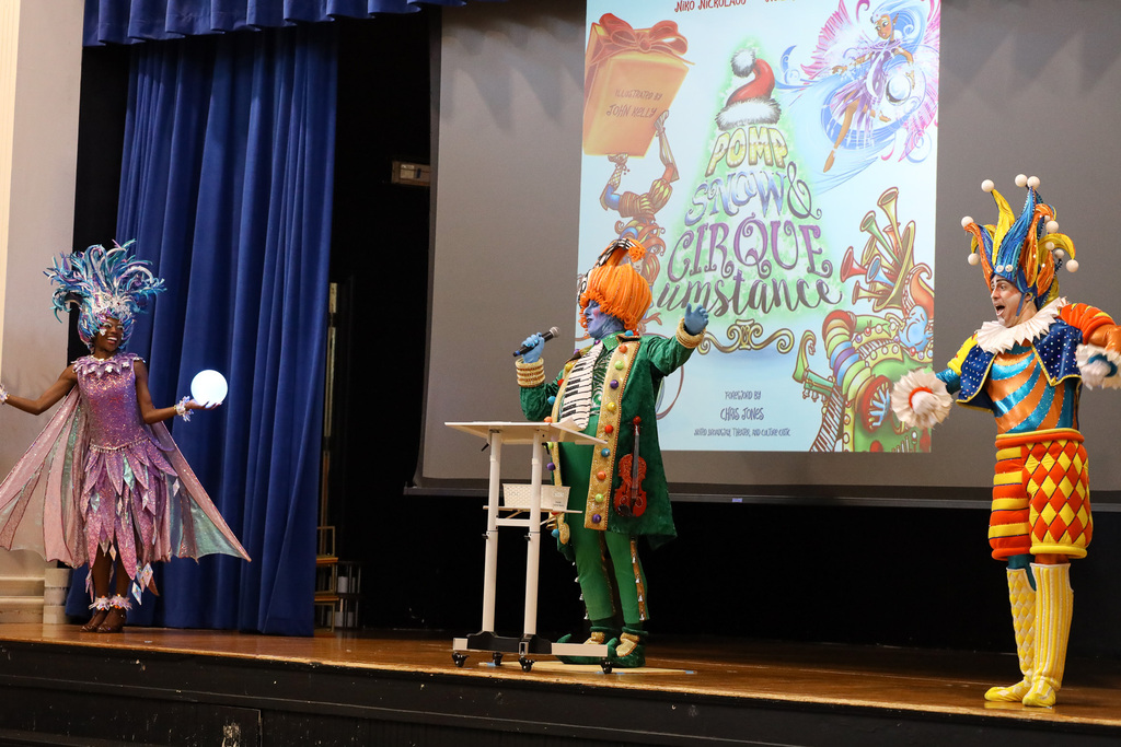 Three people in colorful costumes stand on a stage. One holds a glowing ball, and another appears to speak into a microphone.