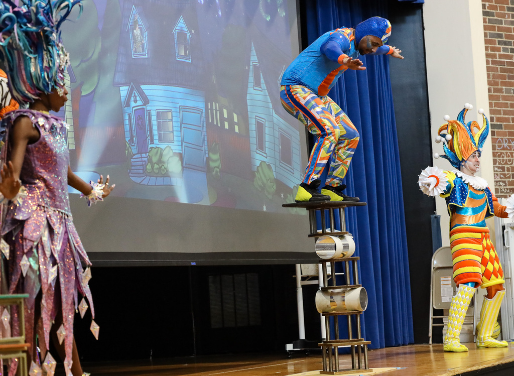 Three people on stage in colorful costumes. One on a platform, another standing, and one sitting on a stack of cans.