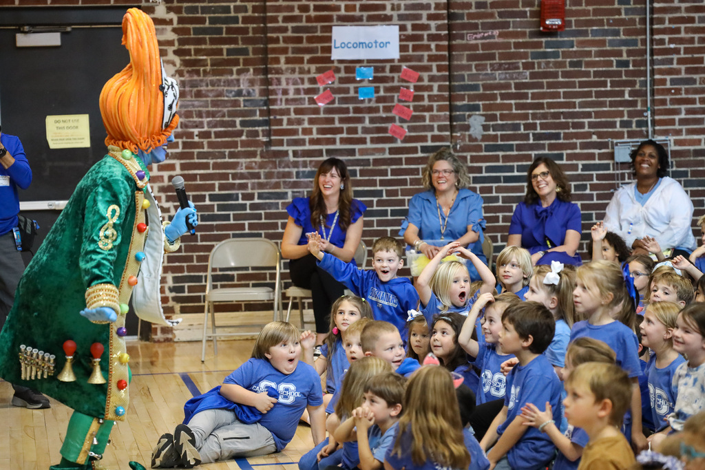 A group of children in blue shirts watch a person in a green clown costume at a school event.