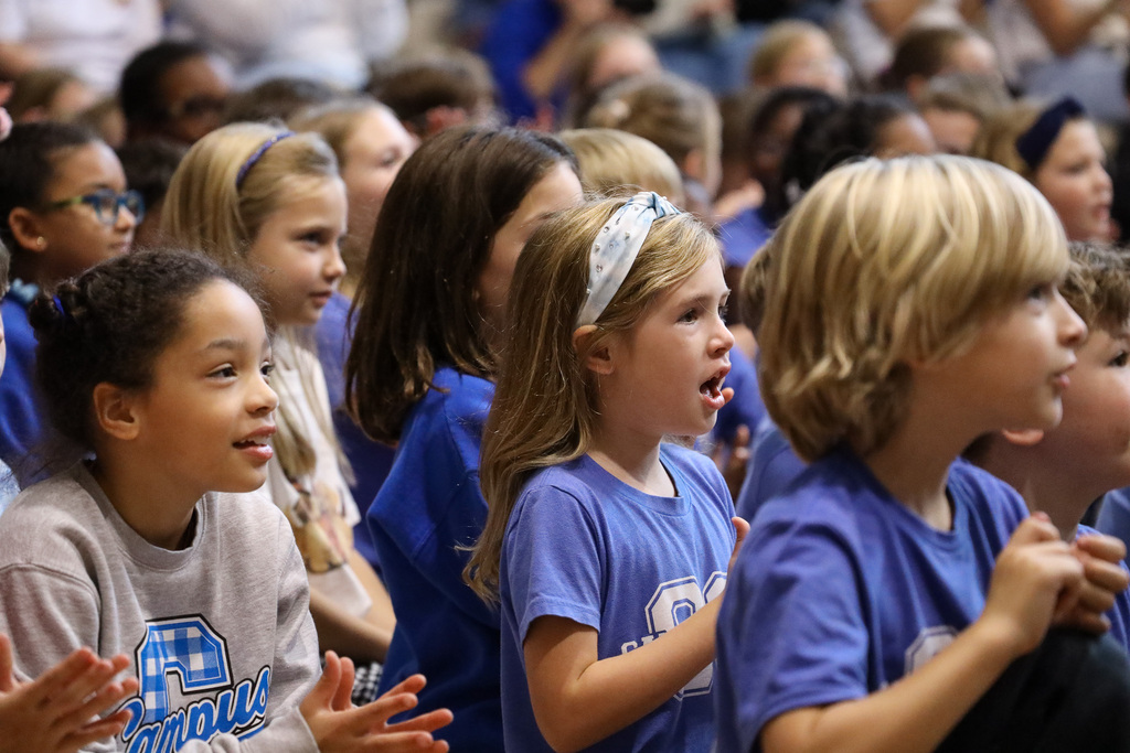 A crowd of students, some wearing blue shirts, sit in a stadium and clap their hands.
