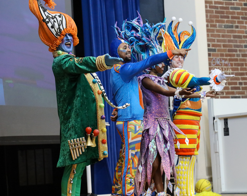 Four adults on stage in colorful costumes, holding props and playing instruments. Blue curtain and brick wall in background.