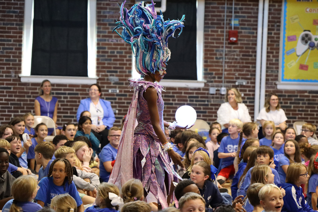 Large crowd of children seated in front of a woman wearing a tall, multicolored headdress.
