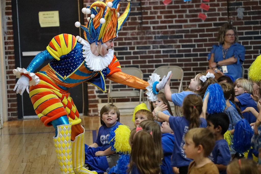 A person in a colorful clown costume entertains a group of children seated in a circle.
