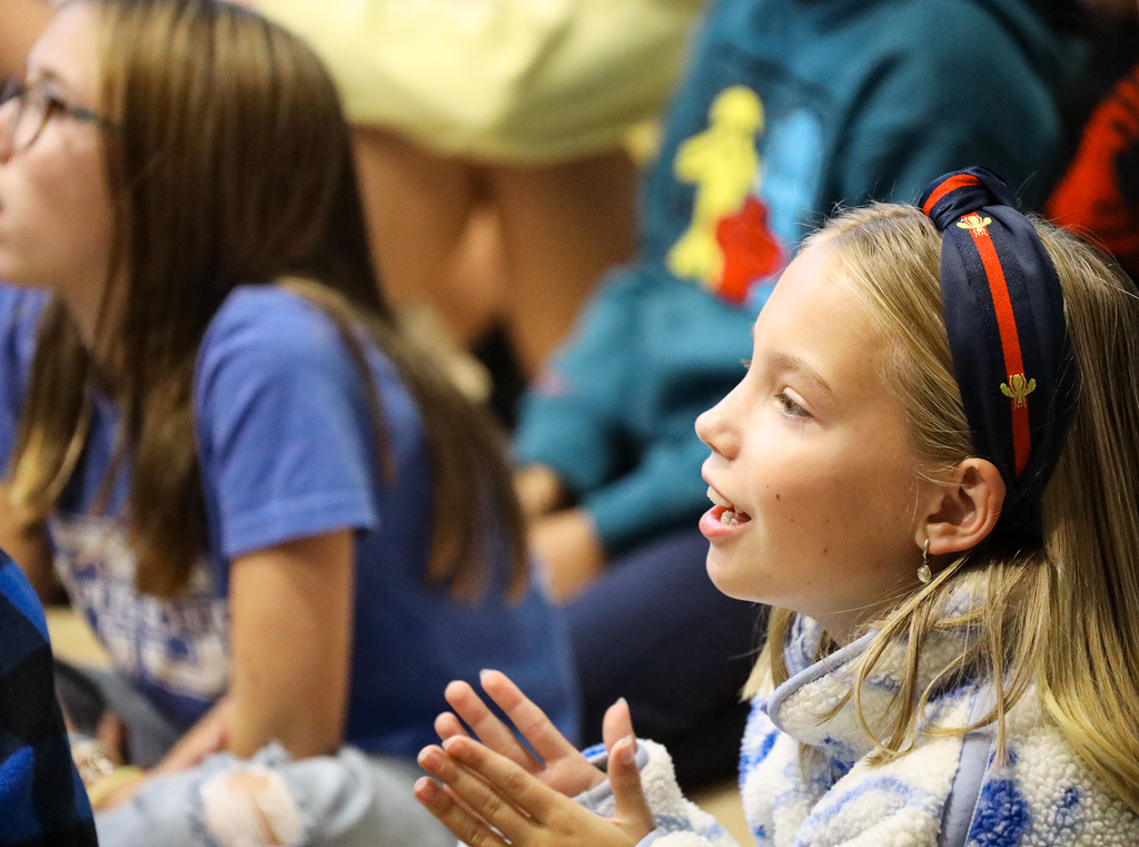 Several students seated, one girl with a headband clapping and looking up, another girl with glasses seated next.
