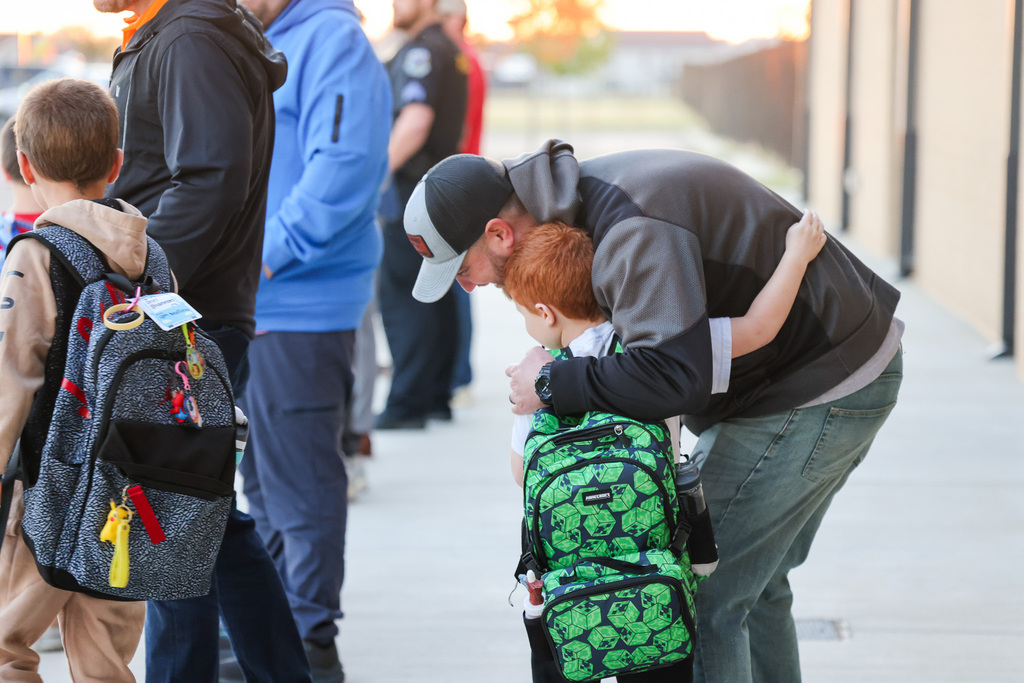 Several people wait in line on a sidewalk. A man hugs a young boy in front of him.