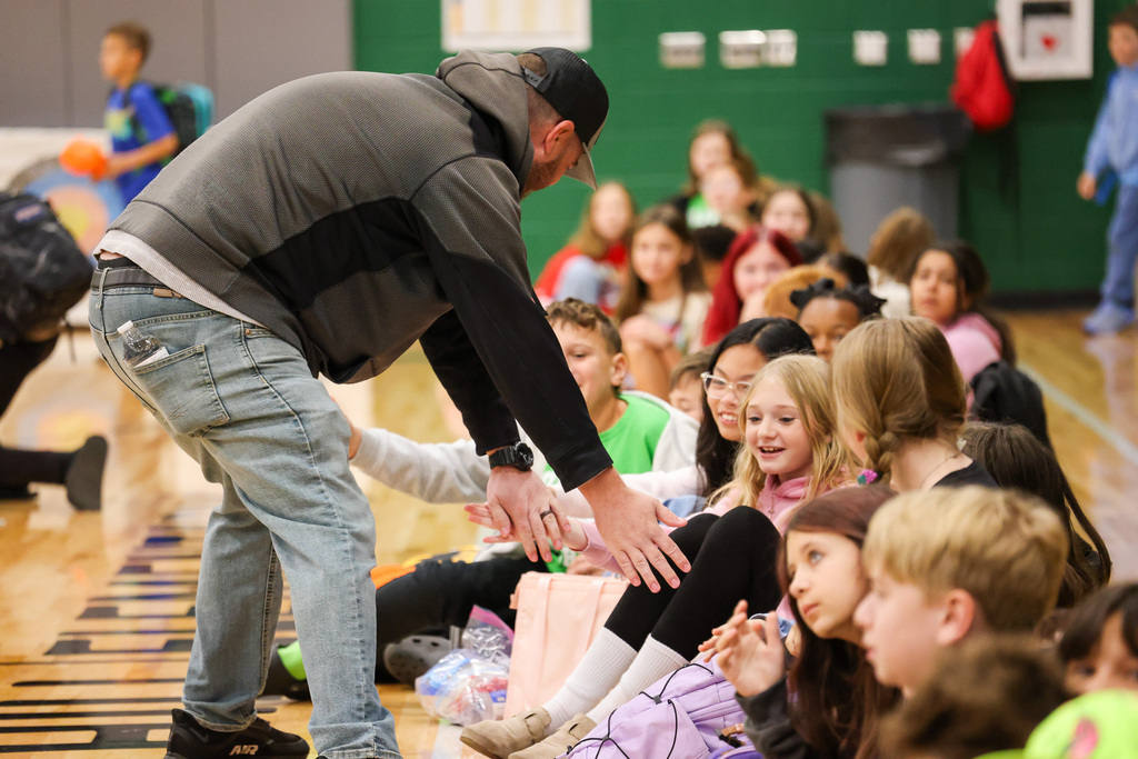 Man standing with group of seated children, hands on knees, children smiling, looking at man.