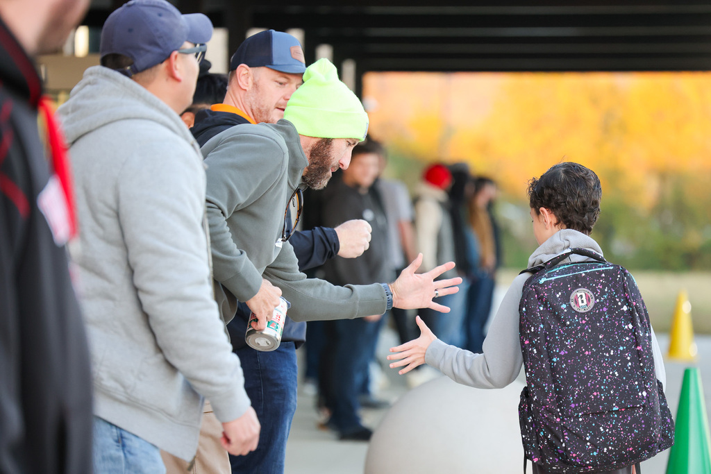 Five people stand outside a building. A person wearing a backpack greets a man with a handshake.