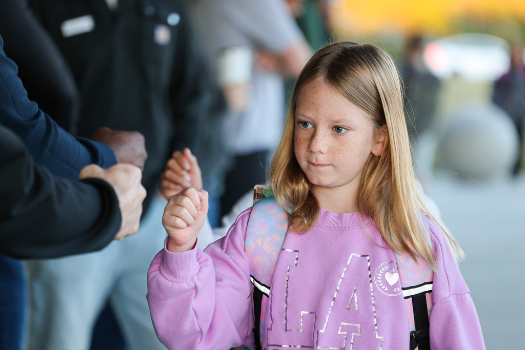 A young girl with blond hair walks with a person holding her hand. They are surrounded by other people.