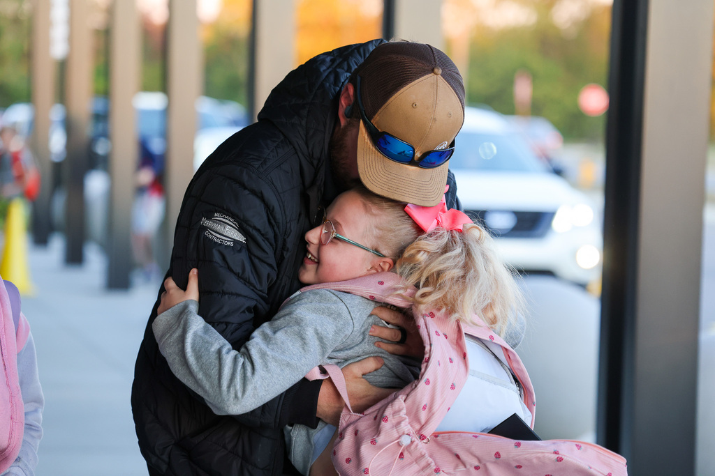 A man hugs a girl in glasses and a pink bow, standing under a building with metal columns.