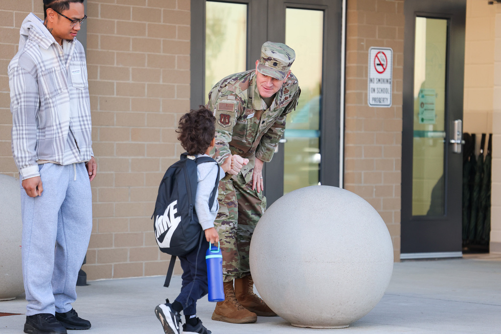 A military man in uniform helps a child with a backpack by a large white sphere. An adult stands nearby.