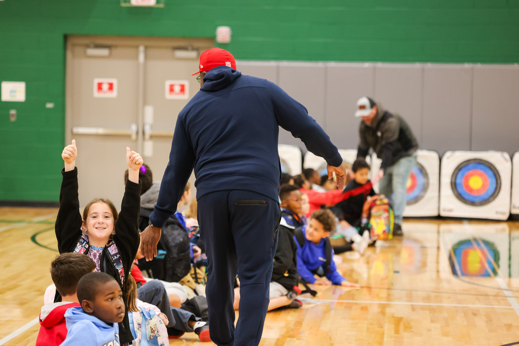 Group of children seated on a gym floor with a man walking past them toward an archery target.