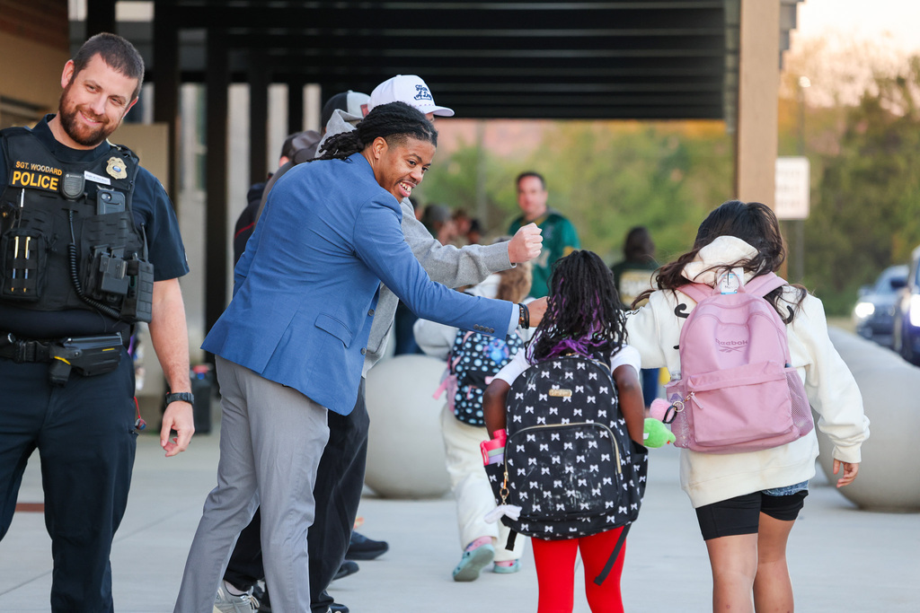 Man in blue suit high-fives child with black backpack. Two other children with backpacks walk behind.
