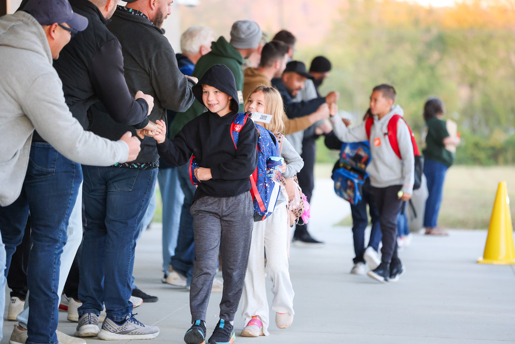 Students and adults in line on a sidewalk with trees in the background. Some students carry backpacks.