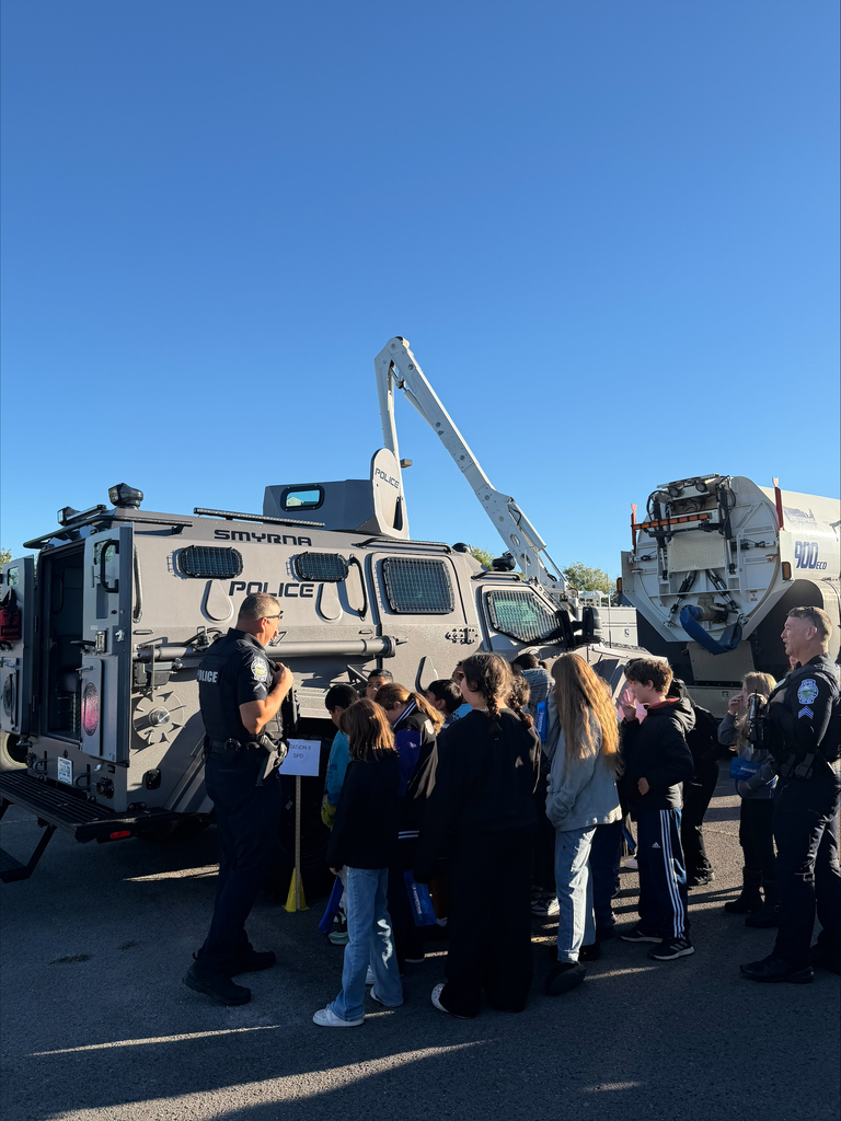 Police officer stands before group of students, two large armored vehicles in background, clear blue sky.