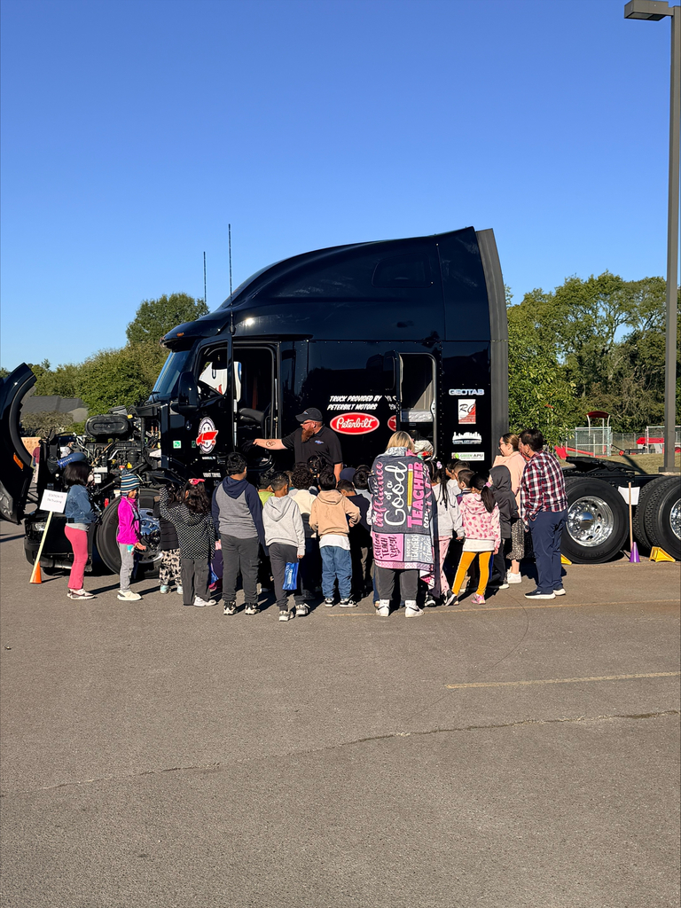 Students gather around a large semi-truck with open hood, near a traffic cone.