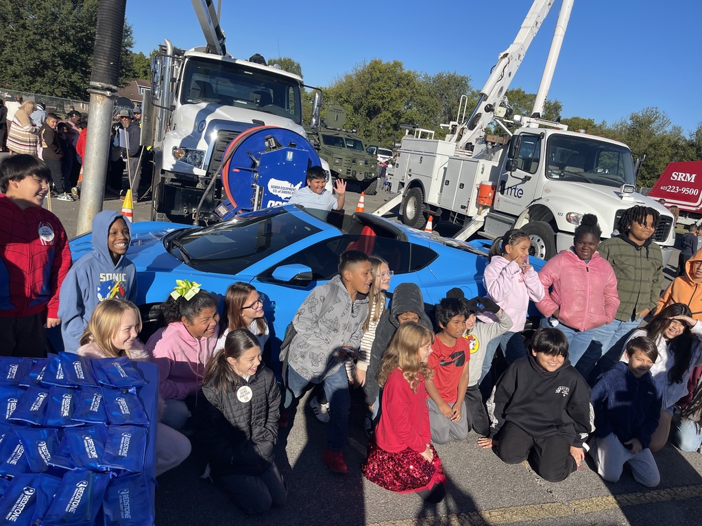 Students and adults stand near a blue sports car and trucks; a pile of plastic bags sits to the left.