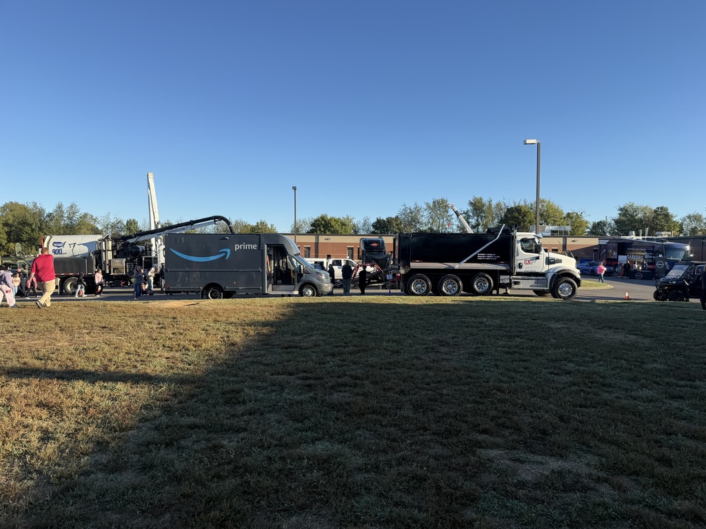 Several vehicles parked on grass. People standing near them. Street lights, trees, and a blue sky.