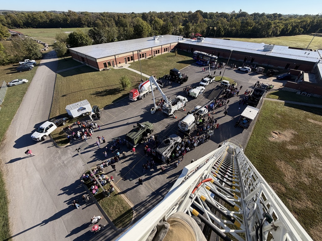 Aerial view of school grounds with vehicles parked in front of a building. Ladders and people are present.