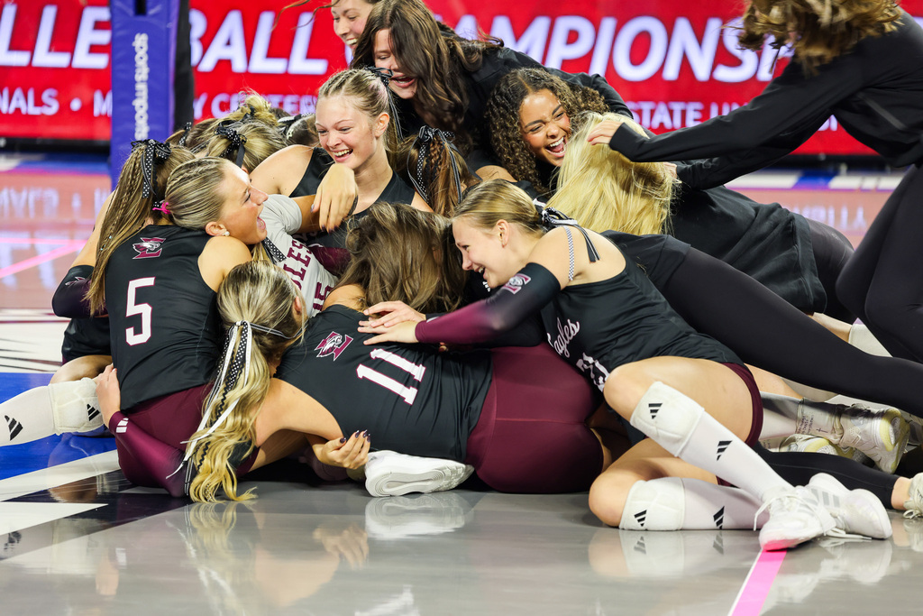Girls volleyball team celebrates on the floor with red and white stripes, wearing maroon and black uniforms.