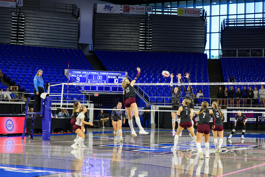 Volleyball players in maroon uniforms jumping to hit a ball over a net, a scoreboard in the background.