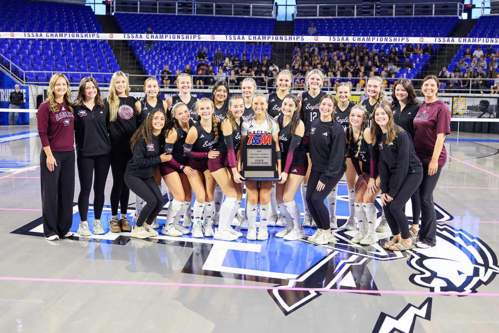 Team with trophy on a court; people in maroon, black, and white jerseys; blue bleachers in background.