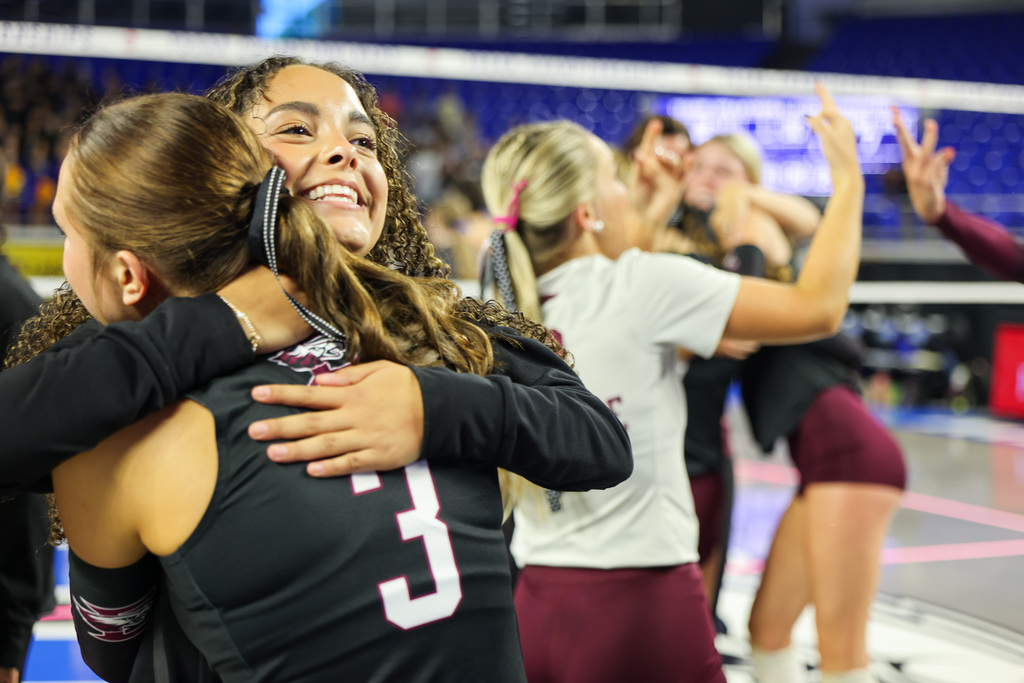 Two girls hugging in a sports uniform with the number 3. Other girls with ponytails standing behind them.
